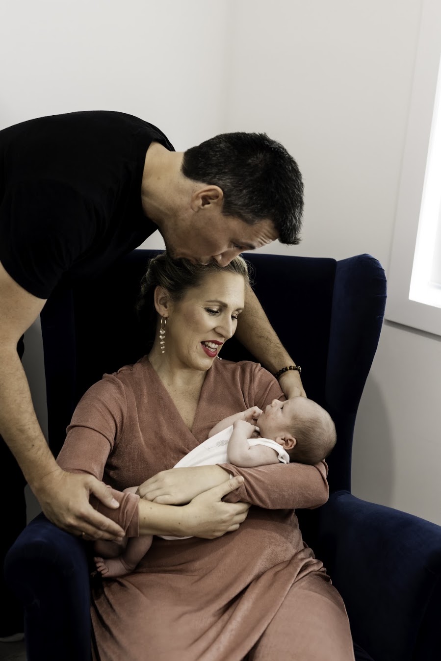A father kisses his wife's head as she cradles their newborn in a navy armchair